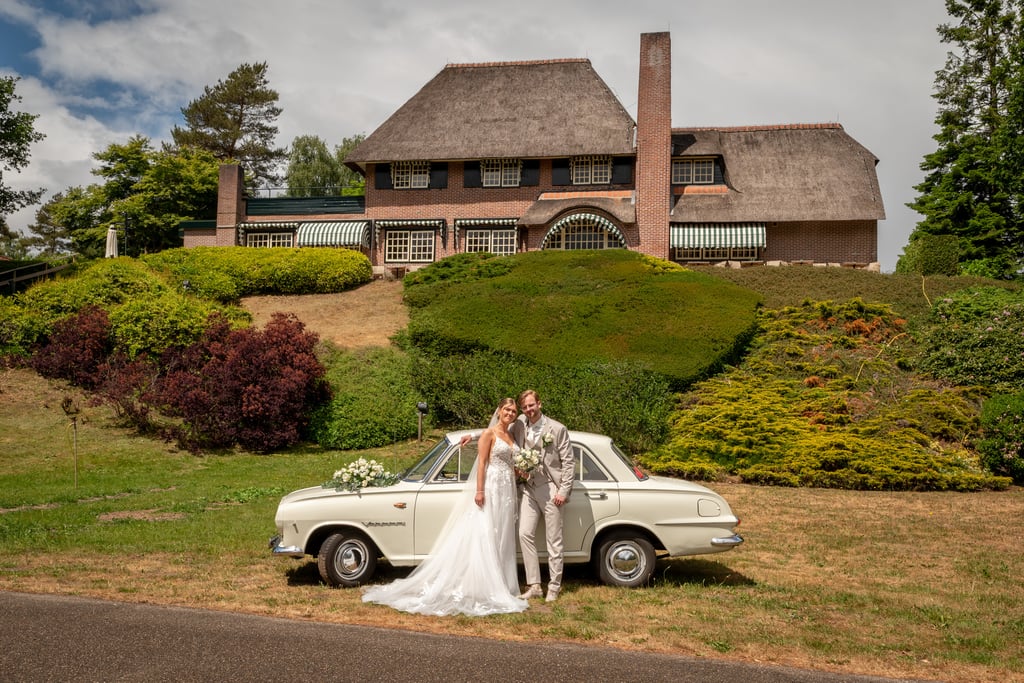 Bruidspaar bij hun klassieke trouwauto voor het huis
